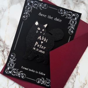 May include: A black save-the-date card with a decorative white border and a black cat silhouette. The card includes the names "Abbi" and "Peter" with a heart, and the date "10.31.2025". The card is inside a burgundy envelope.