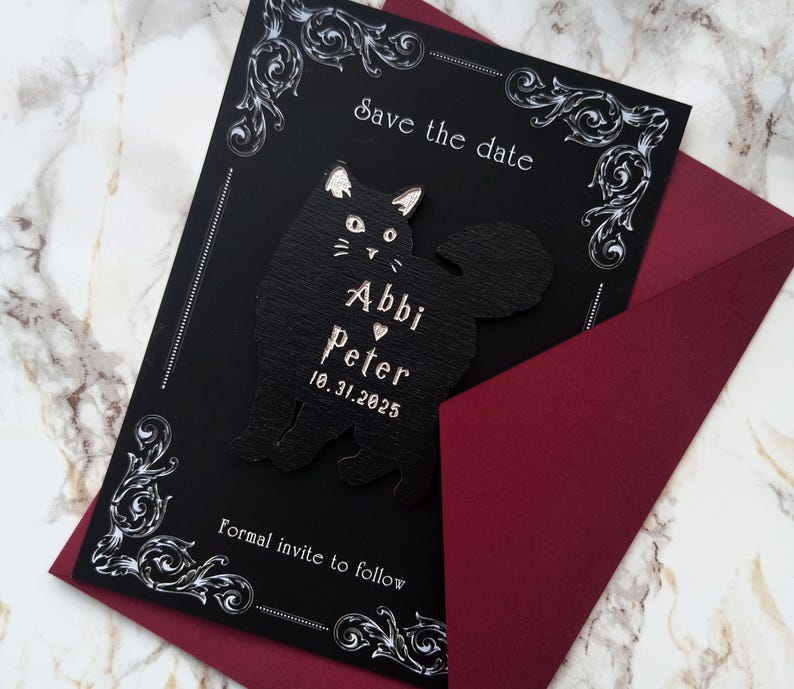 May include: A black save-the-date card with a decorative white border and a black cat silhouette. The card includes the names "Abbi" and "Peter" with a heart, and the date "10.31.2025". The card is inside a burgundy envelope.