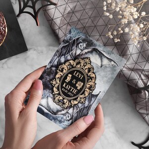 Puede incluir: Una invitaci&oacute;n de boda negra y dorada con un dise&ntilde;o g&oacute;tico que presenta murci&eacute;lagos y un marco decorativo con el texto "Levi & Kaytie 04.01.2023".