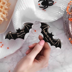 May include: Two black bat-shaped wedding favours with gold lettering. The first bat says "Mr. & Mrs. Deacon 11.16.21". The second bat says "Brittany & Michael 10.16.21".