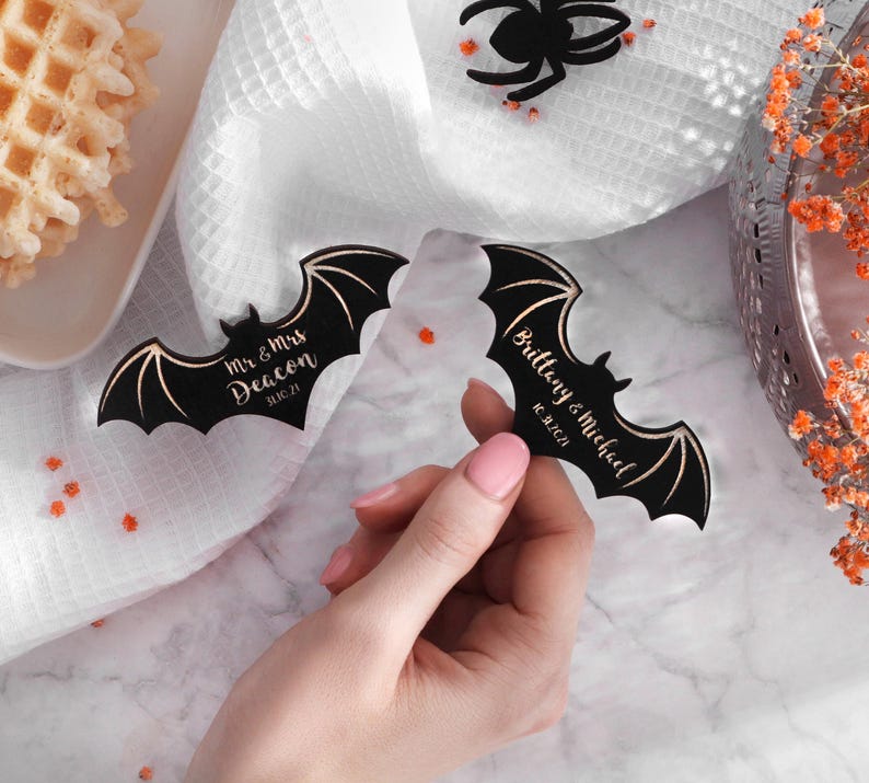 May include: Two black bat-shaped wedding favours with gold lettering. The first bat says "Mr. & Mrs. Deacon 11.16.21". The second bat says "Brittany & Michael 10.16.21".