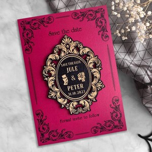 Puede incluir: Una tarjeta roja de guardar la fecha con bordes negros ornamentados y un marco dorado ornamentado con el texto "Save the Date Jule & Peter 31.10.2023" en su interior. El texto "Formal invite to follow" est&aacute; en la parte inferior de la tarjeta.