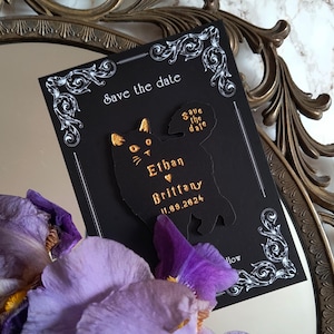 May include: Black "Save the Date" card with a cat silhouette, gold lettering, and decorative white flourishes. The card includes the names "Ethan" and "Brittany" and the date 11.09.2024. Purple flowers and a decorative mirror frame are in the background.