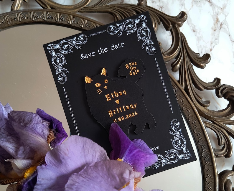 May include: Black "Save the Date" card with a cat silhouette, gold lettering, and decorative white flourishes. The card includes the names "Ethan" and "Brittany" and the date 11.09.2024. Purple flowers and a decorative mirror frame are in the background.