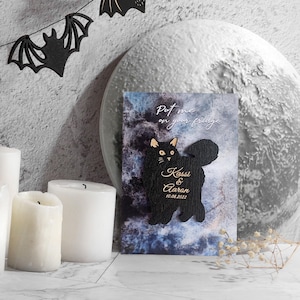 May include: A black cat-shaped magnet with the text "Put me on your fridge" and the names "Kassi & Aaron" with the date "10.08.2022". The magnet is set against a blue and gray background, with decorative candles and bat-shaped cutouts.