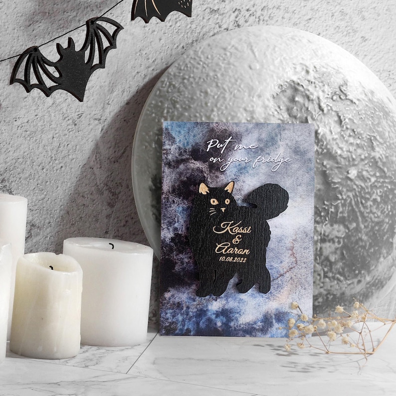 May include: A black cat-shaped magnet with the text "Put me on your fridge" and the names "Kassi & Aaron" with the date "10.08.2022". The magnet is set against a blue and gray background, with decorative candles and bat-shaped cutouts.