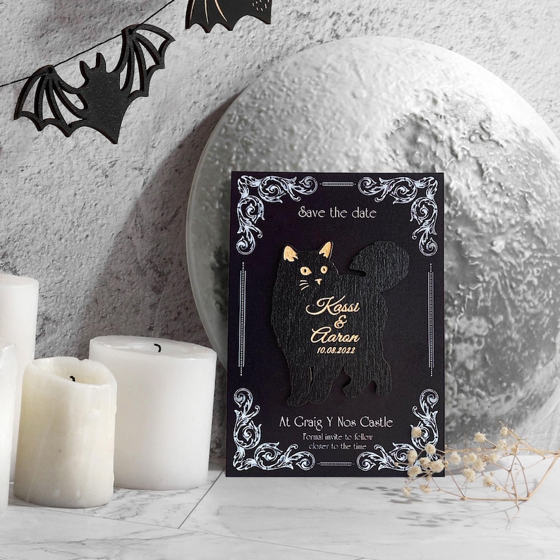 May include: A black wedding invitation card with a cat silhouette and gold accents. The card reads "Save the date" and includes the names "Kassi & Aaron" and the date "10.08.2022". Decorative elements include white flourishes and a castle location.