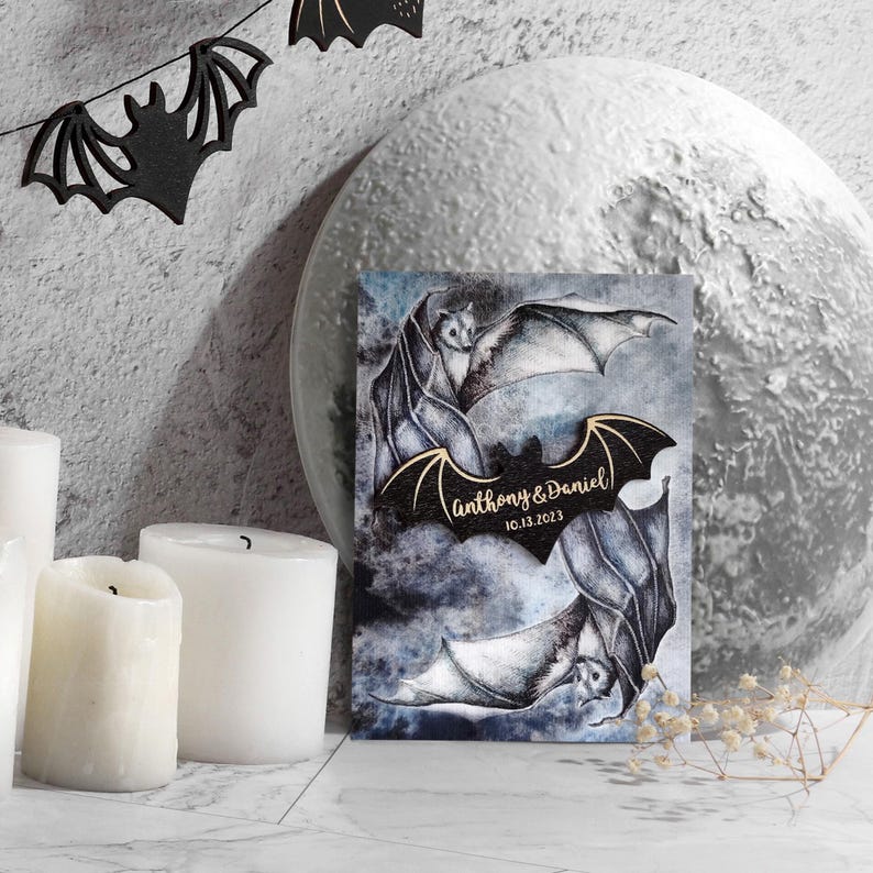 May include: A wedding invitation with a dark blue background featuring two bats with gold wings. The bats are flying in a circle around a moon. The text "Anthony & Daniel 10.13.2023" is written in gold lettering on the bats' wings.