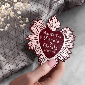 K&ouml;nnte beinhalten: Eine rote, herzf&ouml;rmige Save-the-Date-Karte aus Holz mit goldenem Metallic-Design. Die Karte tr&auml;gt den Text "Save the Date Renata & Gerald 06.08.2024".