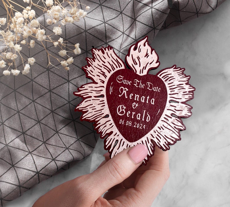 K&ouml;nnte beinhalten: Eine rote, herzf&ouml;rmige Save-the-Date-Karte aus Holz mit goldenem Metallic-Design. Die Karte tr&auml;gt den Text "Save the Date Renata & Gerald 06.08.2024".