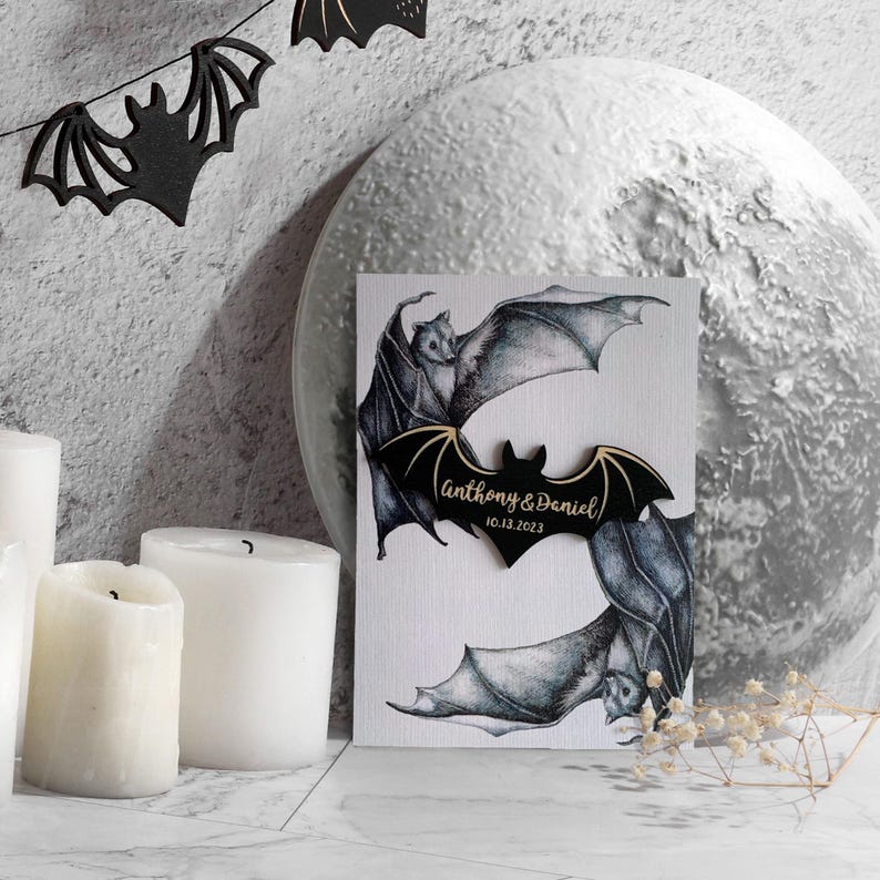 May include: A white card with two black bats drawn on it. The bats are in flight and the card has a wooden bat with the text "Anthony & Daniel 10.13.2023" on it.