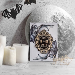 Puede incluir: Una tarjeta de anuncio de boda con un marco adornado en negro y dorado con los nombres Levi & Kaytie y la fecha 04.01.2023. La tarjeta est&aacute; decorada con una ilustraci&oacute;n en blanco y negro de murci&eacute;lagos.