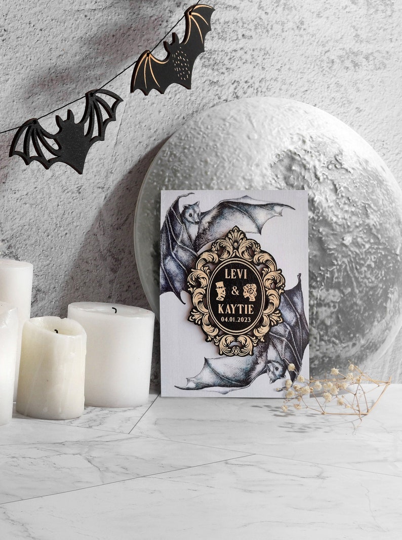 Puede incluir: Una tarjeta de anuncio de boda con un marco adornado en negro y dorado con los nombres Levi & Kaytie y la fecha 04.01.2023. La tarjeta est&aacute; decorada con una ilustraci&oacute;n en blanco y negro de murci&eacute;lagos.