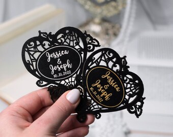 Puede incluir: Dos recuerdos de boda negros cortados con láser en forma de corazón con intrincados diseños de filigrana. Cada recuerdo presenta un óvalo con los nombres "Jessica & Joseph" y la fecha "10.31.2022" en letras blancas y doradas.