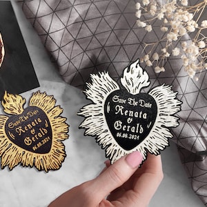 K&ouml;nnte beinhalten: Zwei Save-the-Date-Karten in Herzform mit einer Flamme oben. Die Karten sind schwarz und wei&szlig; mit goldenen Akzenten. Der Text auf den Karten lautet "Save the Date" "Renata & Gerald" und "06.08.2024".