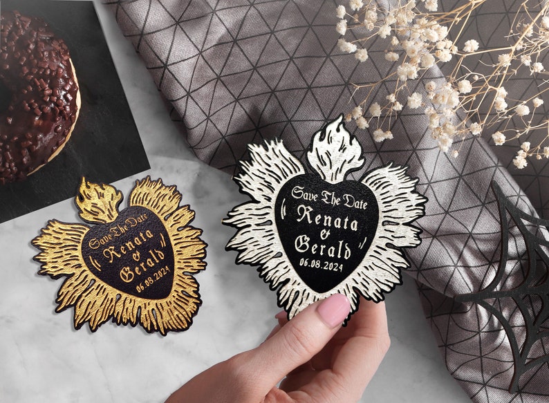 K&ouml;nnte beinhalten: Zwei Save-the-Date-Karten in Herzform mit einer Flamme oben. Die Karten sind schwarz und wei&szlig; mit goldenen Akzenten. Der Text auf den Karten lautet "Save the Date" "Renata & Gerald" und "06.08.2024".