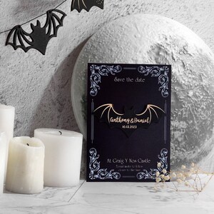 May include: Black save the date card with a white bat design and the text "Save the date", "Anthony & Daniel", "10.13.2023", "At Graig Y Nos Castle", "Formal invite to follow closer to the time".