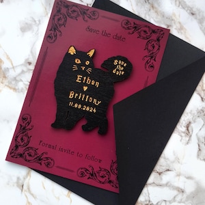 May include: A burgundy save-the-date card with a black cat-shaped embellishment. The cat has gold accents and the names "Ethan" and "Brittony" with the date "11.09.2024". The card is inside a black envelope, with decorative flourishes.