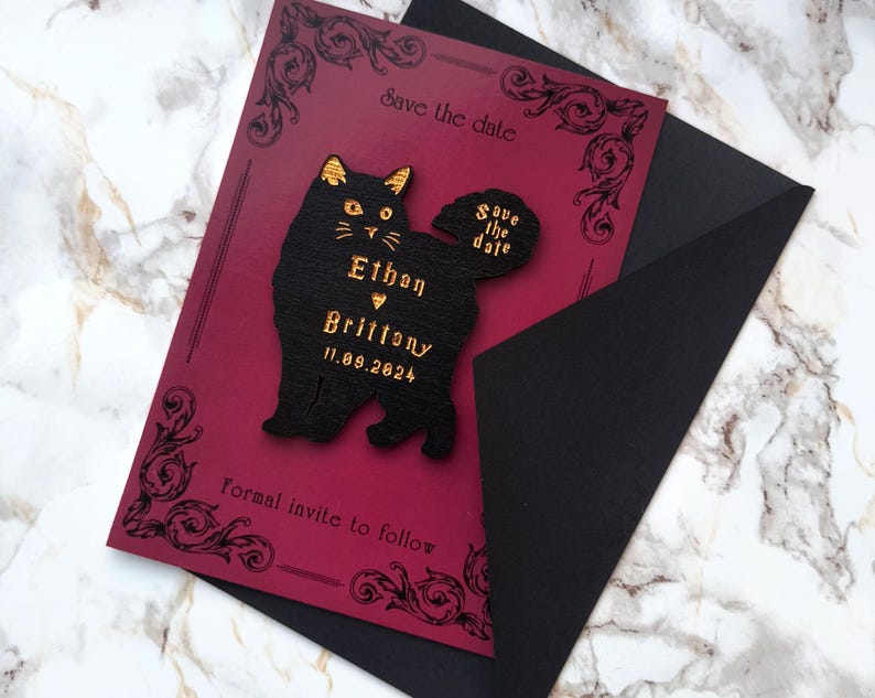 May include: A burgundy save-the-date card with a black cat-shaped embellishment. The cat has gold accents and the names "Ethan" and "Brittony" with the date "11.09.2024". The card is inside a black envelope, with decorative flourishes.