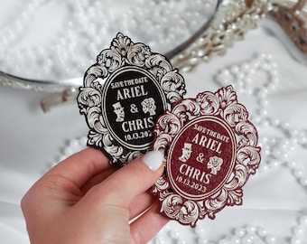May include: Two save the date cards with an ornate frame design. The cards are black and burgundy with white lettering. The text on the cards reads "Save the Date Ariel & Chris 10.13.2023".