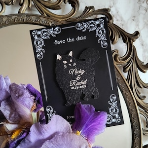 May include: A black "Save the Date" card with a cat-shaped cutout. The card features the names "Nicky & Rachel" and the date "10.26.2024" in white script. The card is decorated with white ornate flourishes.