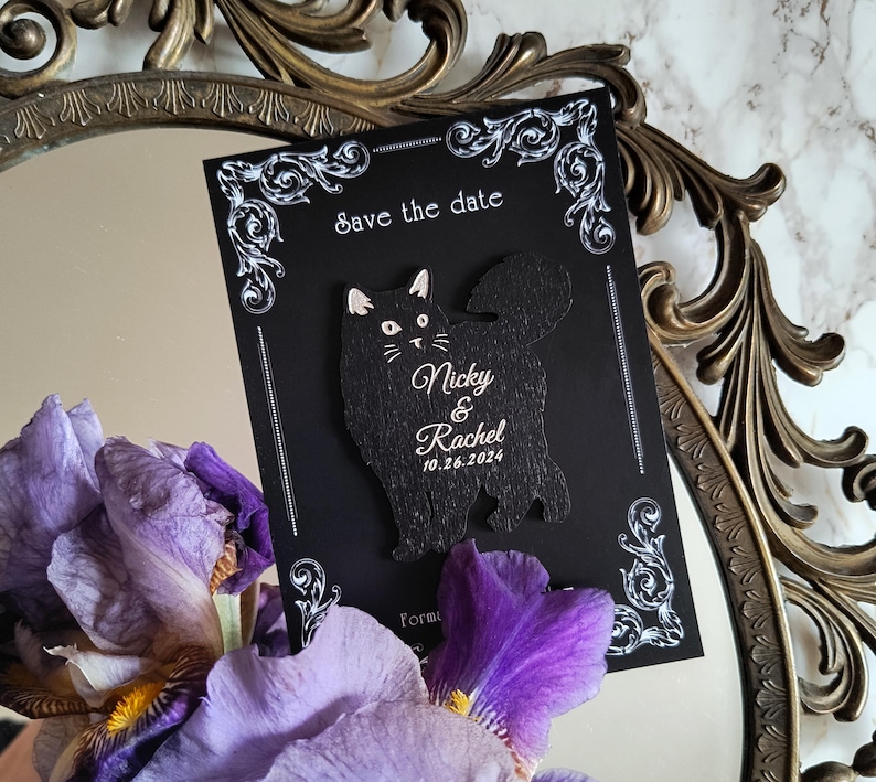 May include: A black "Save the Date" card with a cat-shaped cutout. The card features the names "Nicky & Rachel" and the date "10.26.2024" in white script. The card is decorated with white ornate flourishes.