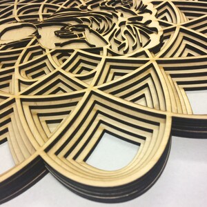 May include: A layered wooden wall art piece with a geometric design. The design is made up of overlapping circles and triangles, creating a complex and intricate pattern. The wood is a light brown color, and the edges of the design are black.