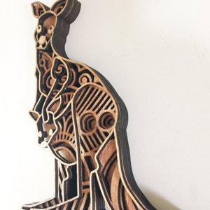 May include: A wooden kangaroo sculpture with intricate laser-cut details. The sculpture is made of multiple layers of wood, creating a 3D effect. The kangaroo is standing on its hind legs, with its tail curled up behind it.