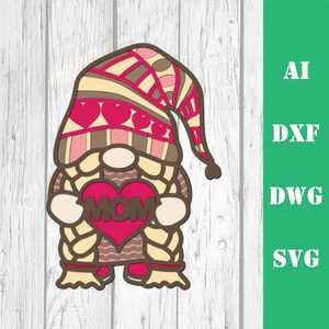 May include: A layered papercraft gnome holding a red heart with the word "MOM" on it. The gnome has a brown hat with pink and brown stripes and a white beard. The gnome is standing on a white wooden background.
