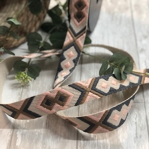 Könnte beinhalten: Gewebtes Band mit einem geometrischen Muster in Rosa, Grau und Braun. Das Band befindet sich auf einer Spule, mit einem neutralfarbenen Hintergrund und Grünzeug.