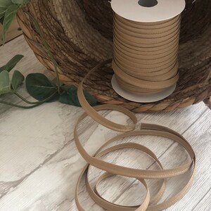 May include: A spool of beige fabric trim with a stitched edge. The trim is wound around a white cardboard spool and is sitting on a woven basket.
