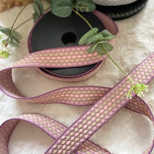 May include: A spool of decorative ribbon in shades of lavender, pink, and gold. The ribbon features a woven pattern of small, diamond-shaped designs. Additional spools of ribbon in red, gray, and black are visible in the background. The ribbon is ideal for crafts and gift wrapping.