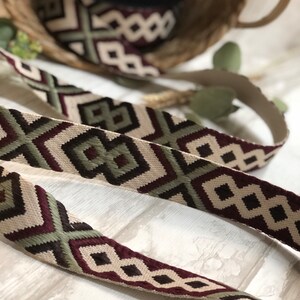 May include: Woven ribbon with a geometric pattern in cream, burgundy, and olive green. The ribbon is displayed in a basket and on a light-colored surface, showcasing its intricate design. This decorative ribbon is suitable for various craft projects.