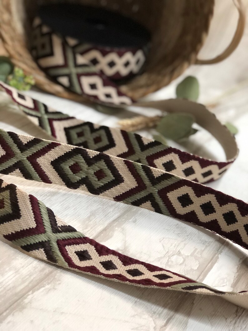 May include: Woven ribbon with a geometric pattern in cream, burgundy, and olive green. The ribbon is displayed in a basket and on a light-colored surface, showcasing its intricate design. This decorative ribbon is suitable for various craft projects.