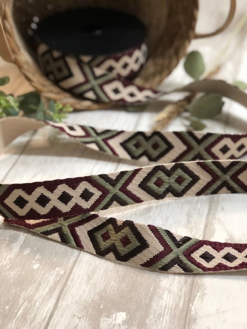 May include: Decorative ribbon with a geometric pattern in burgundy, olive green, and cream. The ribbon is displayed in a roll and in a length, showcasing the repeating design. The ribbon is likely used for crafts or embellishments.