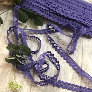 May include: A roll of purple lace ribbon with a scalloped edge and openwork design. The ribbon is partially unrolled, with a sprig of greenery in the foreground. The lace is a vibrant shade of purple.