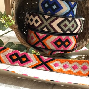 May include: A woven basket holds spools of patterned ribbon in various colors. The ribbons feature geometric designs in black, orange, pink, blue, and beige. A length of the pink and orange ribbon lies in front.