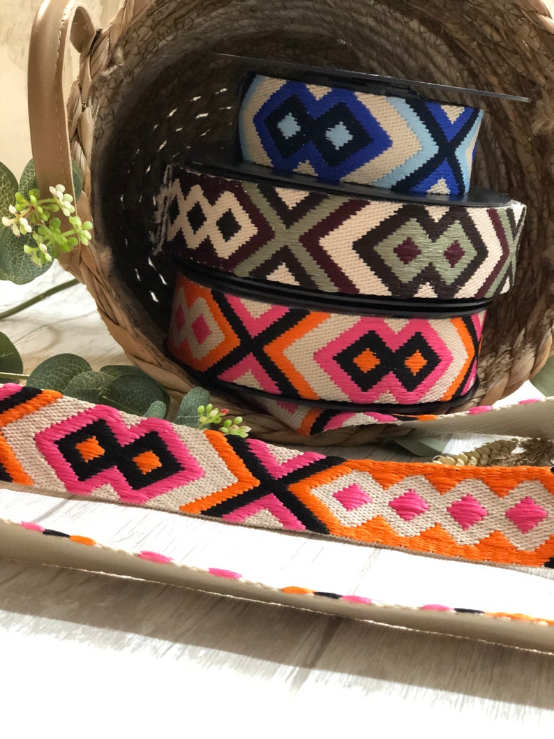May include: A woven basket holds spools of patterned ribbon in various colors. The ribbons feature geometric designs in black, orange, pink, blue, and beige. A length of the pink and orange ribbon lies in front.