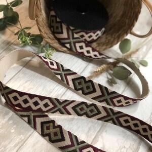 May include: Woven strap with a geometric pattern in burgundy, olive green, and cream. The strap is displayed with a woven basket and a roll of black material. The strap's design features diamond and arrow shapes.