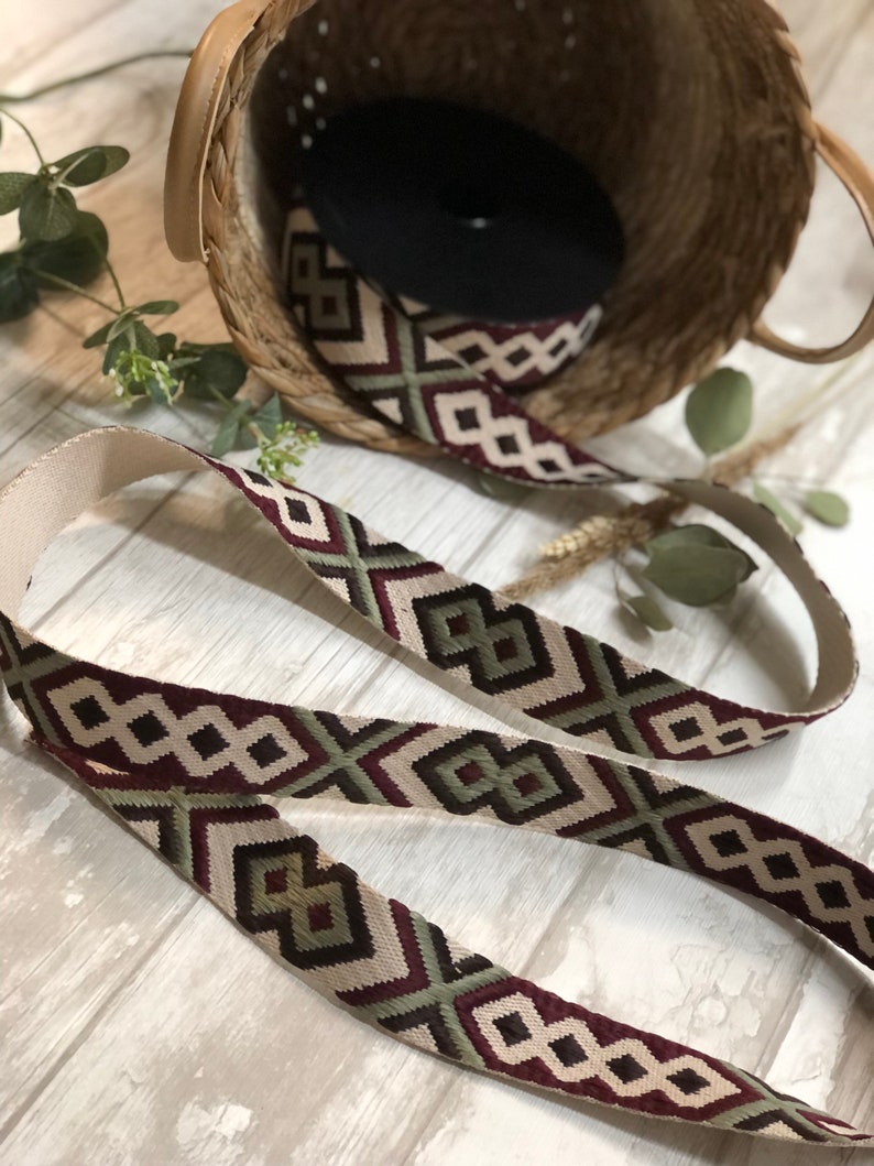 May include: Woven strap with a geometric pattern in burgundy, olive green, and cream. The strap is displayed with a woven basket and a roll of black material. The strap's design features diamond and arrow shapes.