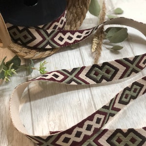 May include: A spool of patterned ribbon with a geometric design in burgundy, olive green, and cream. The ribbon is displayed with a length of the same pattern, showcasing its texture and design. The ribbon is suitable for crafts and decorative projects.