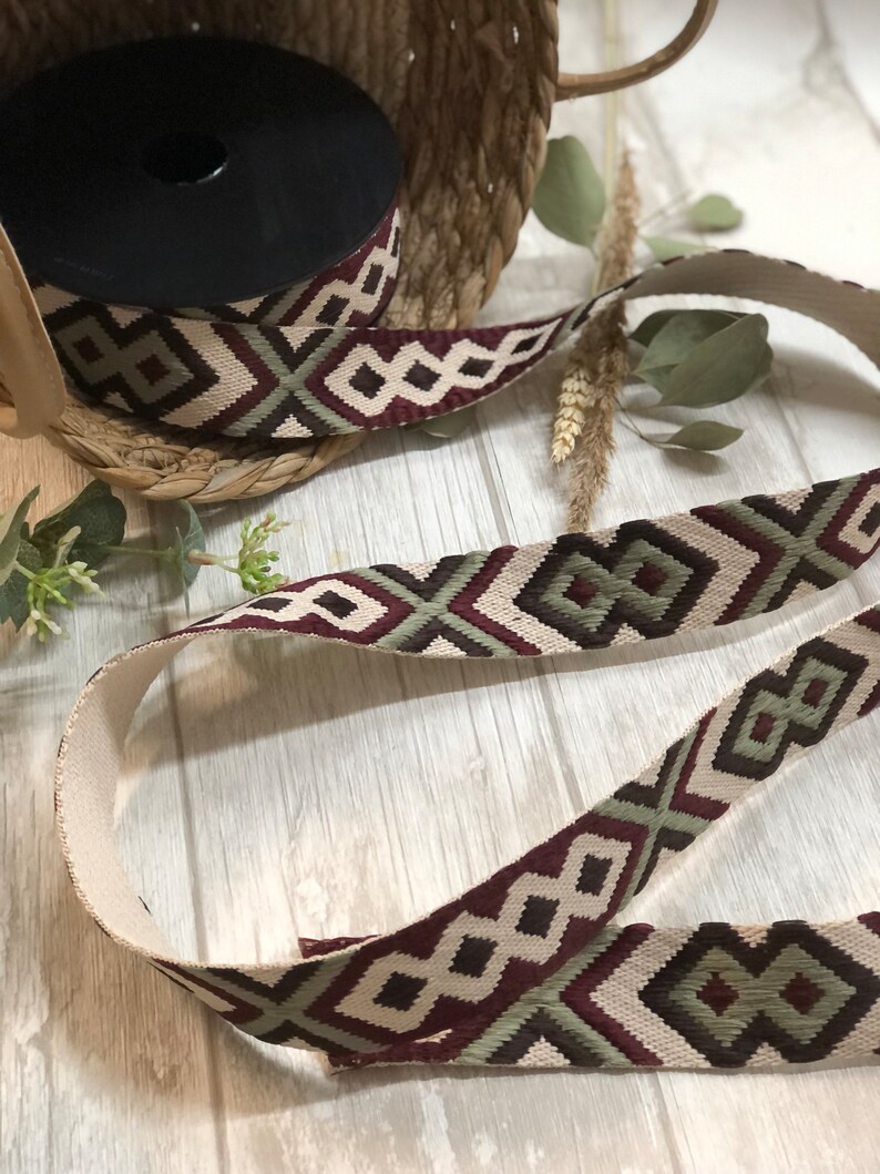 May include: A spool of patterned ribbon with a geometric design in burgundy, olive green, and cream. The ribbon is displayed with a length of the same pattern, showcasing its texture and design. The ribbon is suitable for crafts and decorative projects.