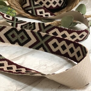 May include: Woven ribbon with a geometric diamond pattern in cream, burgundy, and olive green. A roll of the ribbon sits in a woven basket, with a length of the ribbon unrolled in the foreground. The ribbon is suitable for crafts and sewing projects.