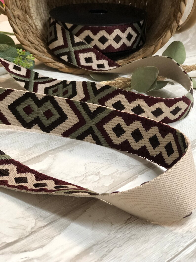 May include: Woven ribbon with a geometric diamond pattern in cream, burgundy, and olive green. A roll of the ribbon sits in a woven basket, with a length of the ribbon unrolled in the foreground. The ribbon is suitable for crafts and sewing projects.