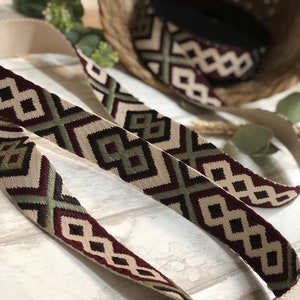 May include: Close-up of a woven strap with a geometric pattern in cream, burgundy, green, and black. The strap is displayed with a roll of matching material in a basket. The strap has a cream-colored backing.