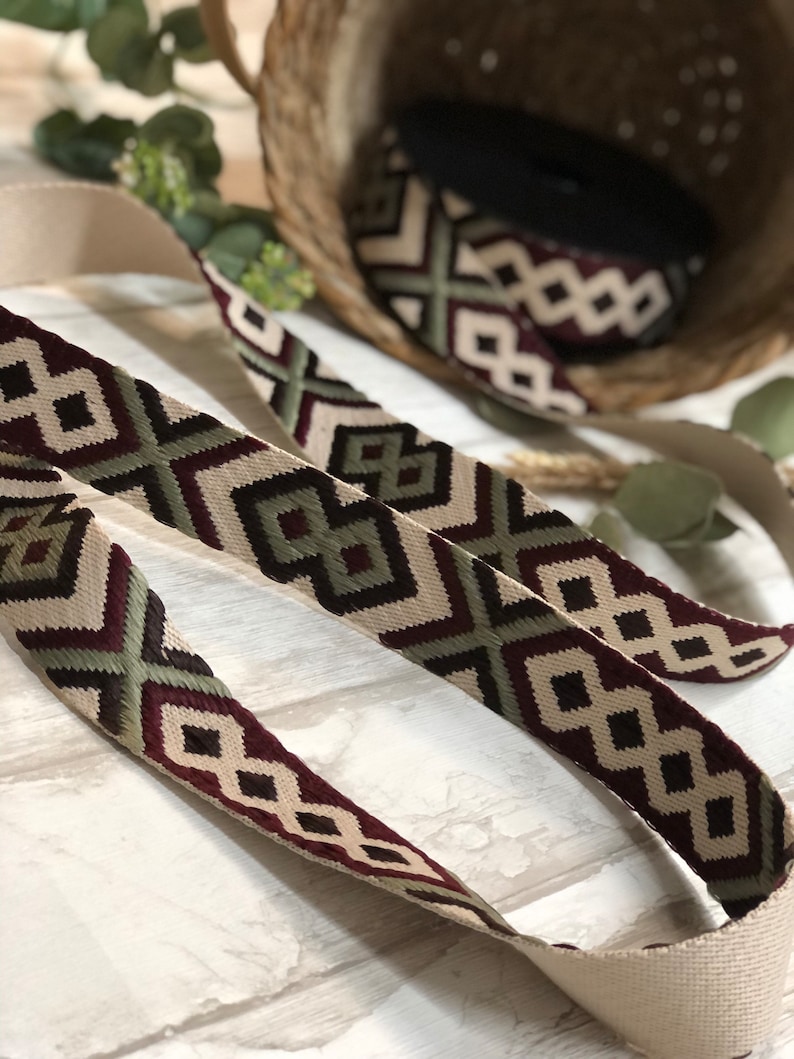 May include: Close-up of a woven strap with a geometric pattern in cream, burgundy, green, and black. The strap is displayed with a roll of matching material in a basket. The strap has a cream-colored backing.
