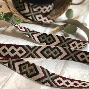 May include: Decorative woven ribbon with a geometric pattern in cream, burgundy, and olive green. A roll of the ribbon sits in a woven basket. The ribbon is ideal for crafting and adding a decorative touch to various projects.