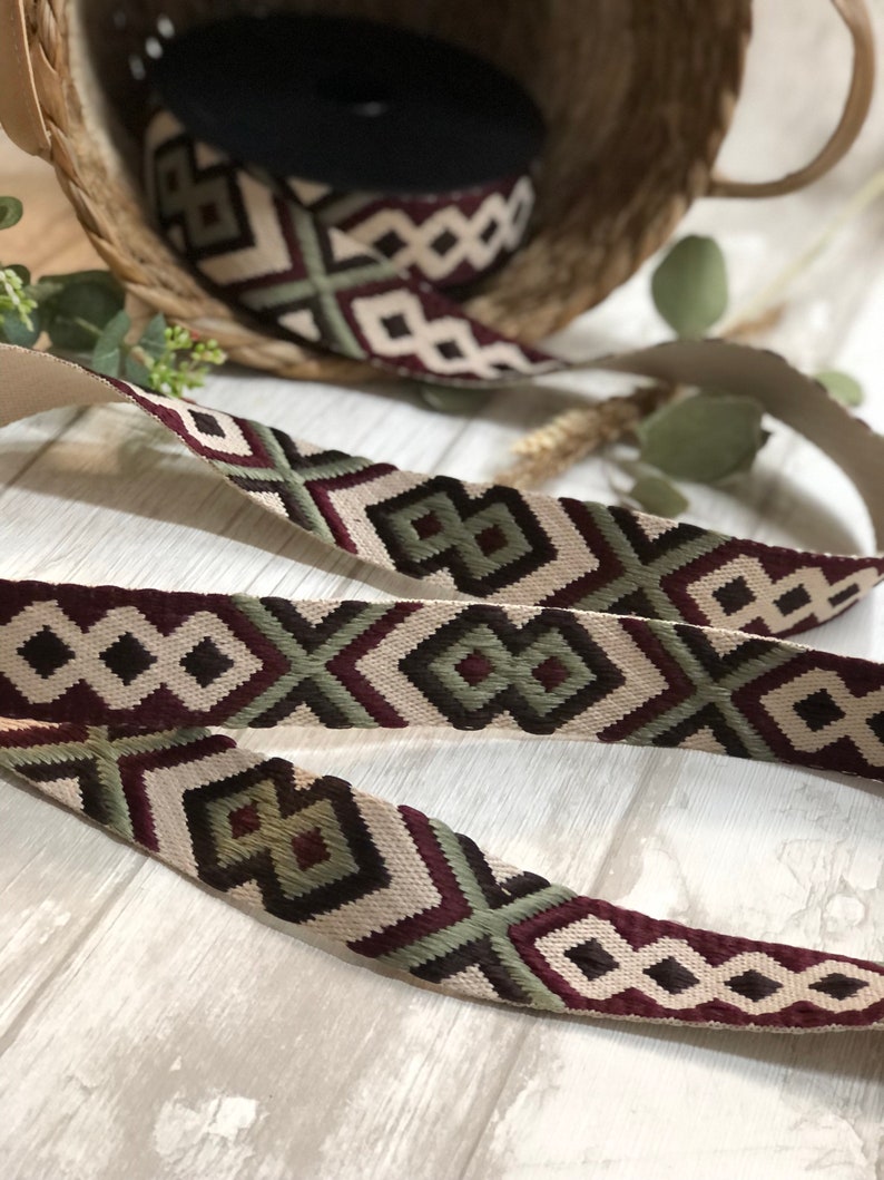 May include: Decorative woven ribbon with a geometric pattern in cream, burgundy, and olive green. A roll of the ribbon sits in a woven basket. The ribbon is ideal for crafting and adding a decorative touch to various projects.
