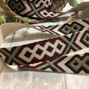 May include: A roll of decorative ribbon with a geometric pattern in cream, burgundy, olive green, and black. The ribbon is displayed on a light wooden surface, with a woven basket in the background.