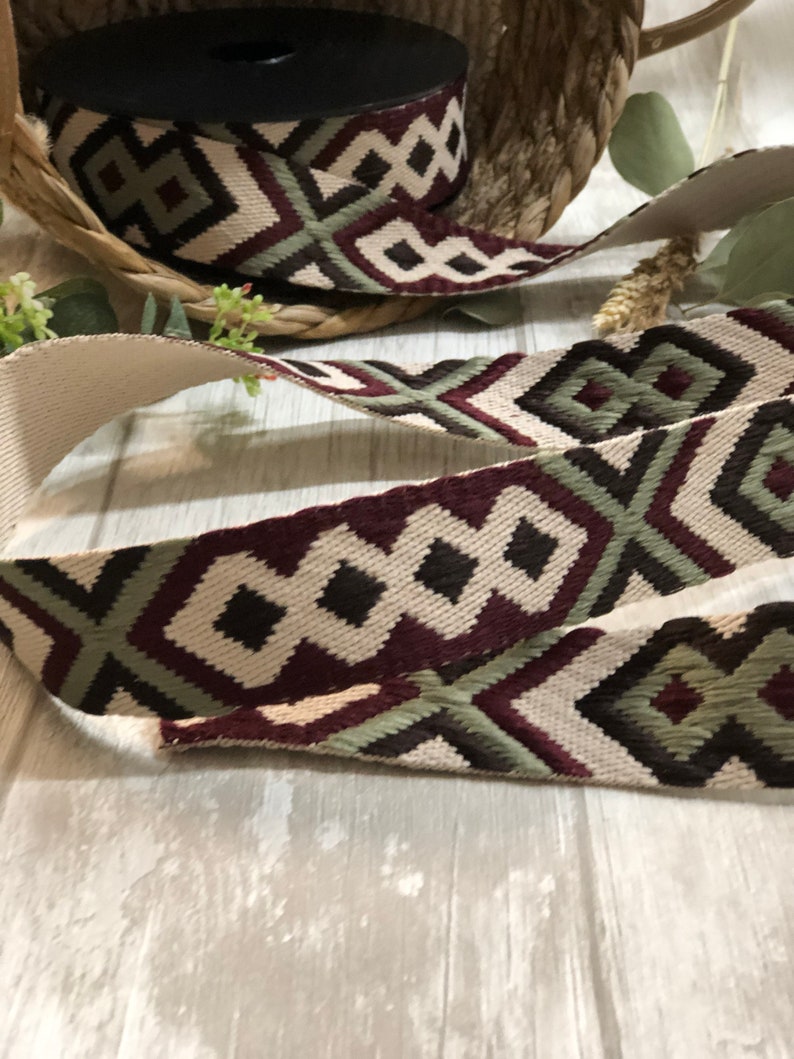 May include: A roll of decorative ribbon with a geometric pattern in cream, burgundy, olive green, and black. The ribbon is displayed on a light wooden surface, with a woven basket in the background.